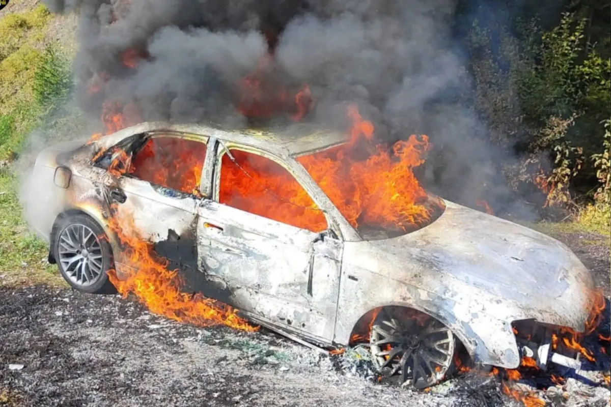 Ein Auto in einer ländlichen Umgebung steht in Flammen. Dichte, schwarze Rauchwolken steigen in die Luft und Flammen durchziehen das Fahrzeug.