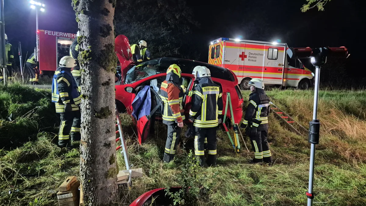 Feuerwehrleute arbeiten an einem roten, verunfallten Auto im Gelände bei Nacht. Ein Rettungswagen vom Deutschen Roten Kreuz steht bereit.