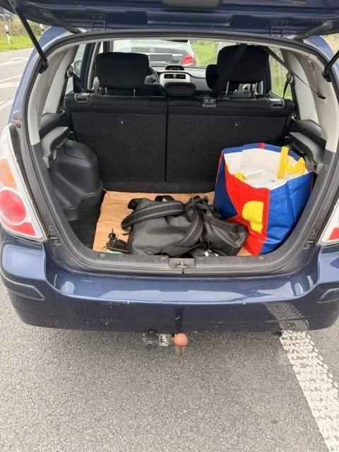 Der Kofferraum eines blauen Autos ist geöffnet und zeigt eine schwarze Tasche sowie eine bunte Einkaufstasche auf einem Holzbrett im Inneren.