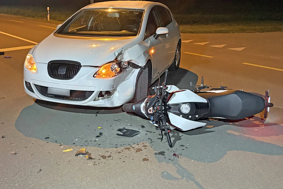 Ein weißes Auto mit sichtbaren Beschädigungen an der Vorderseite steht auf einer Straße. Daneben liegt ein umgefallenes Motorrad ebenfalls auf der Straße.