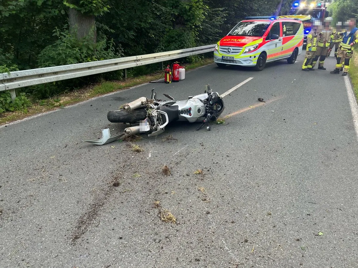 Ein verunfalltes Motorrad liegt auf einer Landstraße, während ein Notarztwagen und Rettungskräfte im Hintergrund zu sehen sind.
