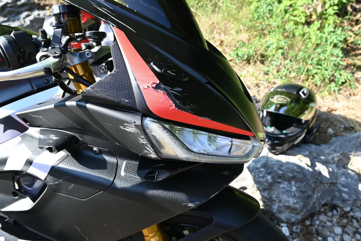 Das Bild zeigt den Vorderteil eines Motorrads mit Kratzern an der Verkleidung. Ein Helm liegt im Hintergrund auf steinigem Boden.