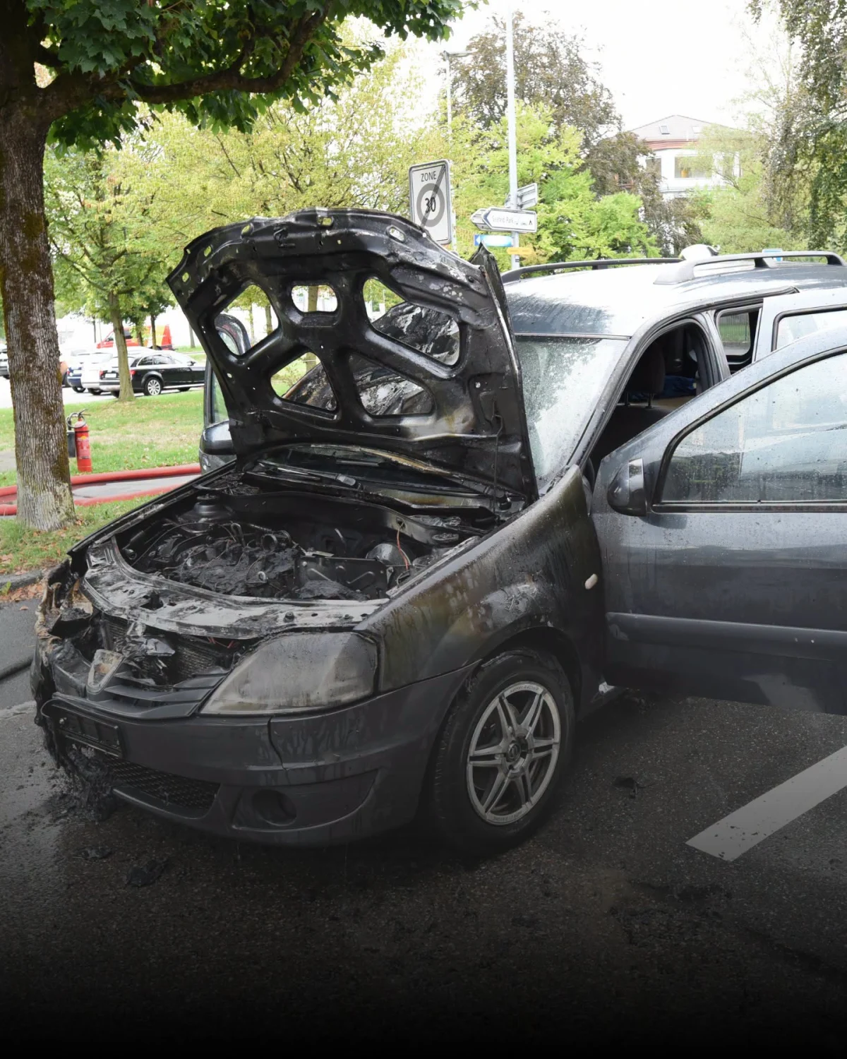 Ein ausgebranntes Auto steht mit geöffneter Motorhaube auf einer Straße. Der Motorraum ist stark beschädigt und verrußt.