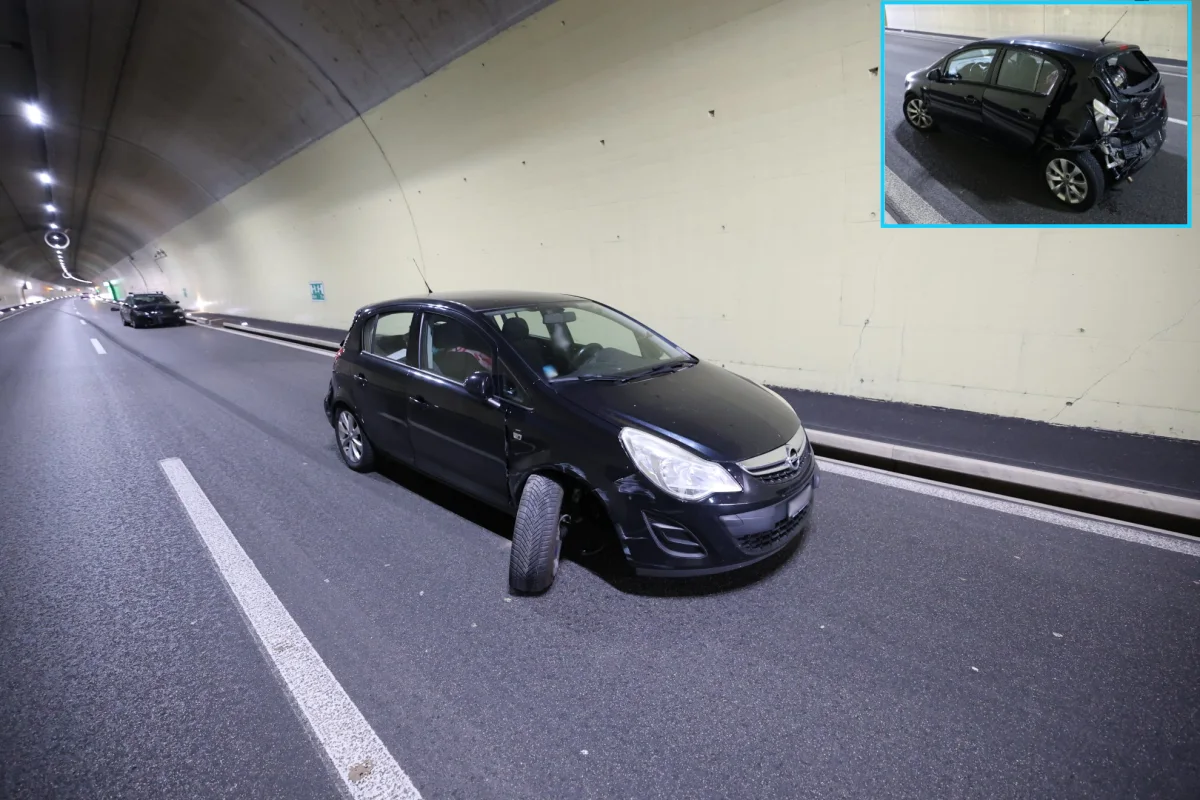 Ein schwarzes Auto mit einem beschädigten Vorderrad steht in einem leeren Tunnel. Im Hintergrund ist ein weiteres Fahrzeug sichtbar, das am Straßenrand parkt.