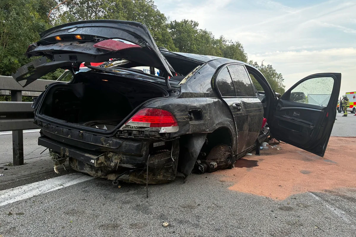 Ein stark beschädigtes Auto steht auf der Autobahn, der Kofferraum und die Türen sind geöffnet. Im Hintergrund ist ein Rettungsfahrzeug zu sehen.