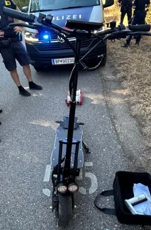 Ein E-Scooter steht auf einer Straße vor einem Polizeifahrzeug. Polizeibeamte sind im Hintergrund sichtbar.