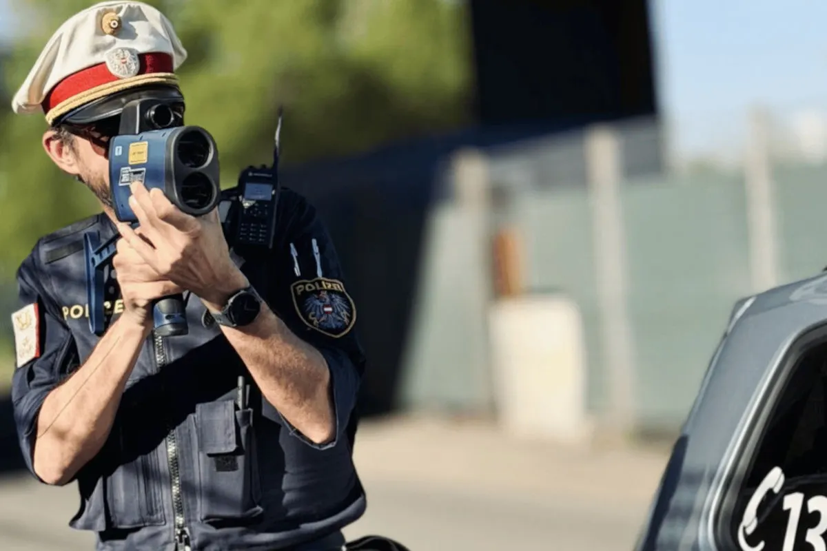 Ein Polizist in dunkler Uniform verwendet ein Geschwindigkeitsmessgerät auf einer Straße. Neben ihm ist ein Teil eines Autos sichtbar.
