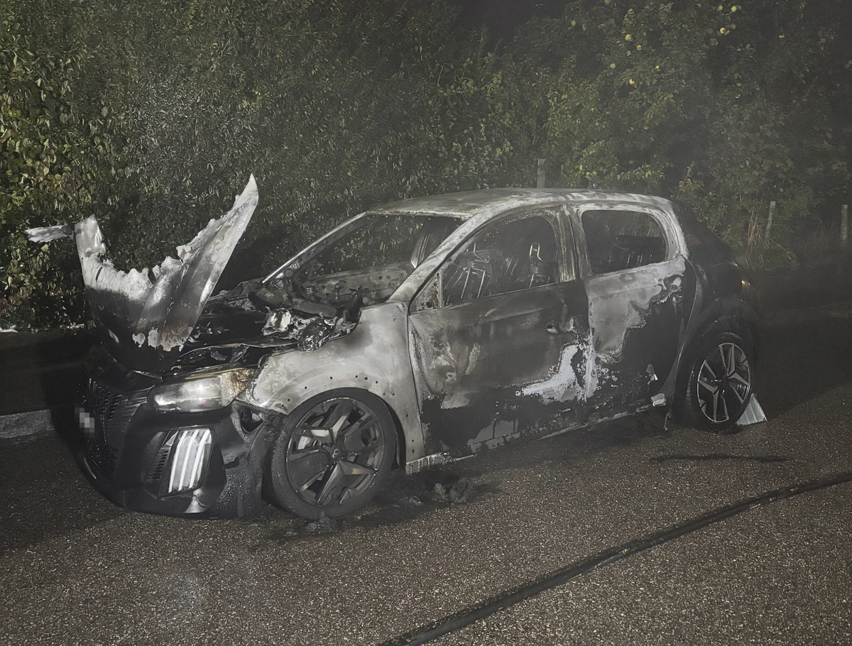 Ein Auto, das auf einer Straße bei Nacht vollständig ausgebrannt ist, die Karosserie ist stark beschädigt, und es sind noch Rauchspuren sichtbar.