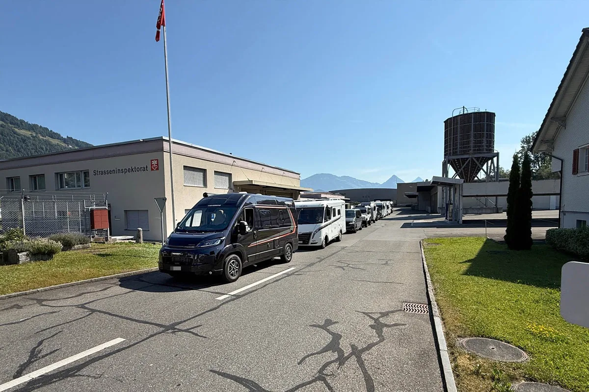 Eine Kolonne von Wohnmobilen steht vor dem Gebäude des Strasseninspektorats. Der Himmel ist klar und die Berge sind im Hintergrund sichtbar.