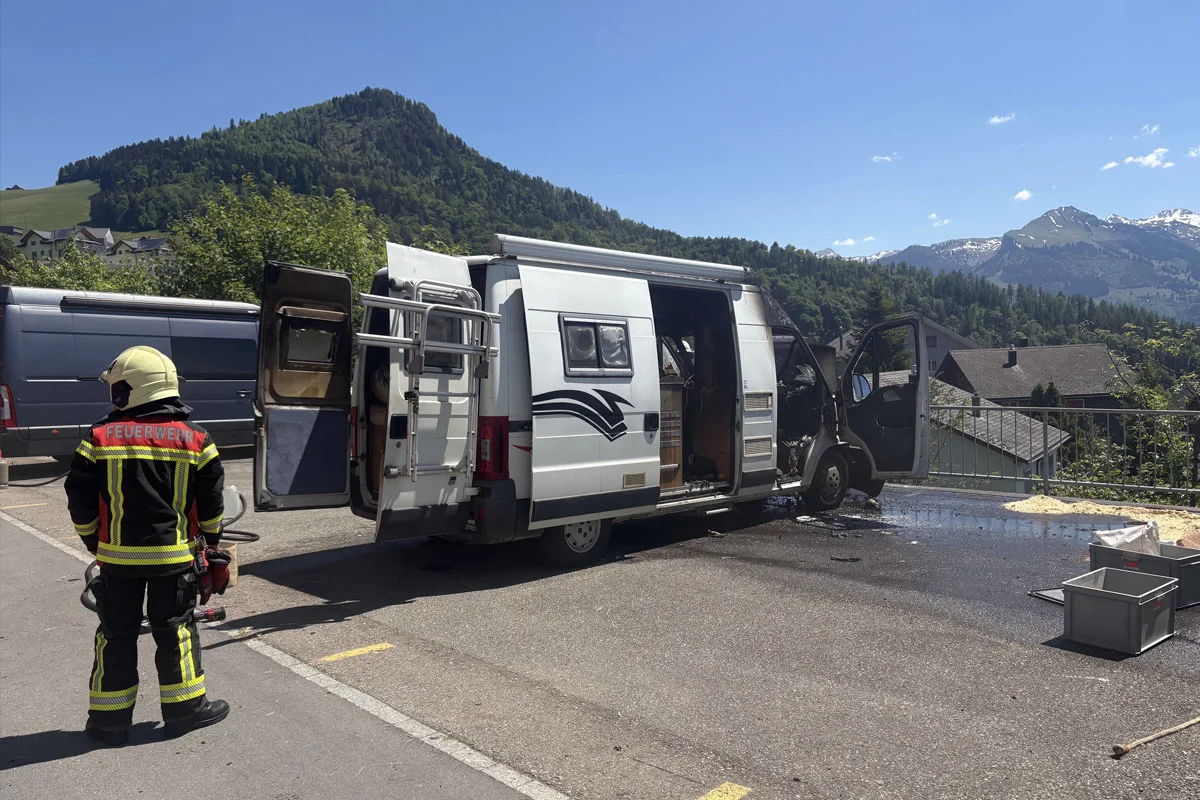 Ein Feuerwehrmann in Schutzkleidung steht auf einem Parkplatz neben einem ausgebrannten weißen Transporter. Im Hintergrund sind Berge und Häuser zu sehen.