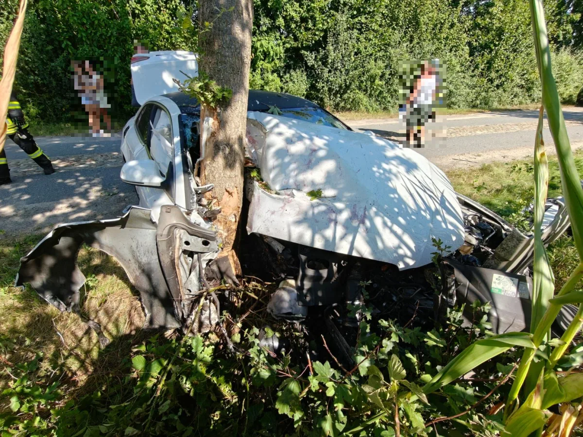 Ein weißes Auto ist frontal gegen einen Baum gefahren, wodurch die Vorderseite stark beschädigt wurde. Eine Person in Feuerwehrkleidung steht links daneben, während mehrere Personen im Hintergrund stehen.