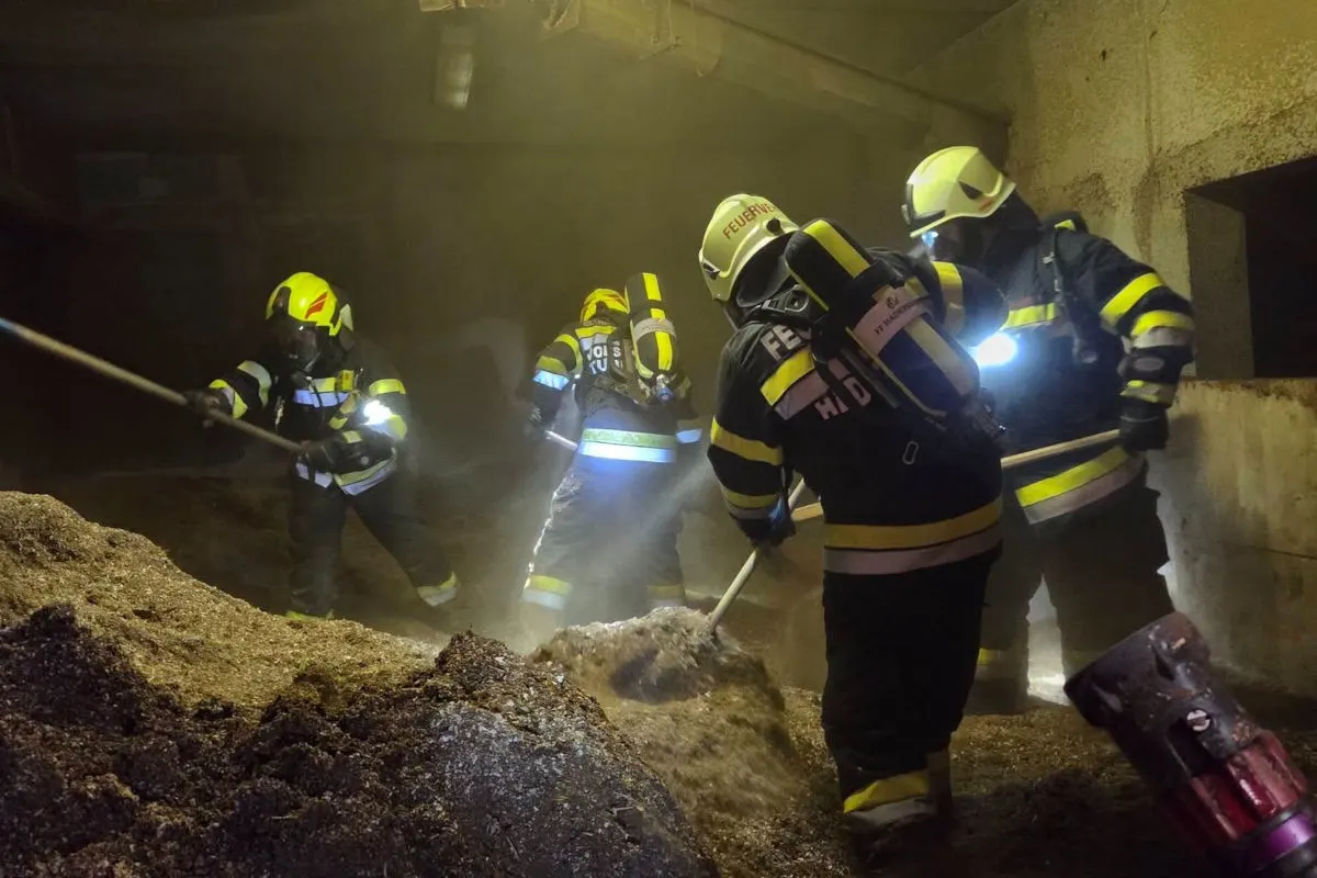 Mehrere Feuerwehrleute mit Atemschutzmasken und Helmen hantieren mit Schaufeln in einem staubigen Raum. Sie tragen Schutzanzüge mit reflektierenden Streifen.
