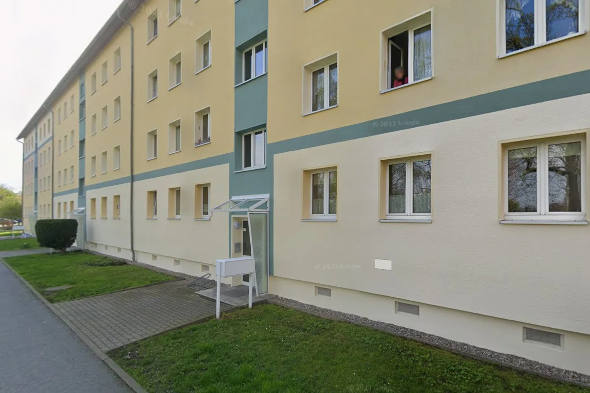 Das Bild zeigt ein mehrstöckiges Wohngebäude mit einer gelben Fassade. Einige Fenster sind geöffnet, und eine Person schaut aus einem Fenster.