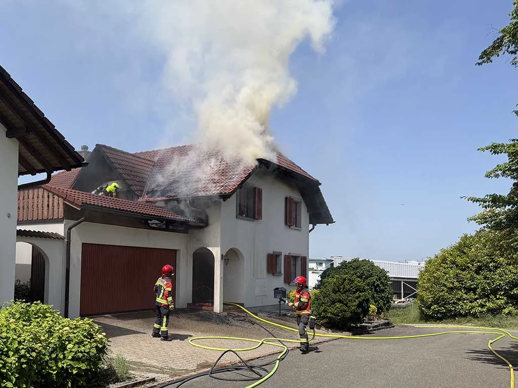 Ein Wohnhaus, aus dessen Dach Rauch aufsteigt, während Feuerwehrleute mit Schläuchen bereitstehen. Der Himmel ist klar.