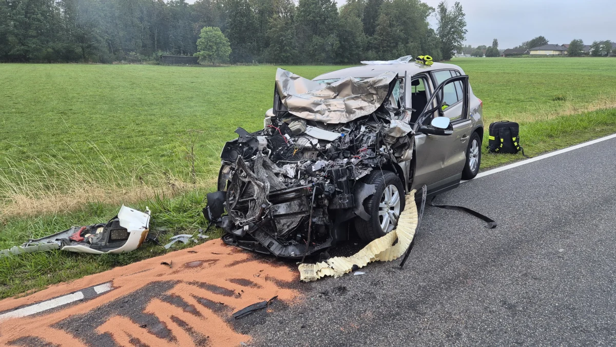 Ein Fahrzeug mit massiven Frontschäden steht auf einer Landstraße. Die Umgebung ist ländlich mit Wiesen und Bäumen.