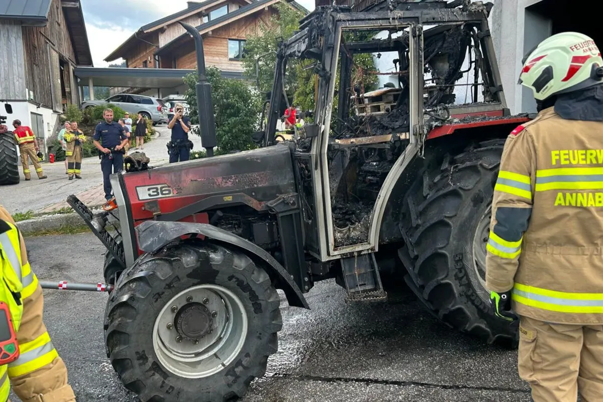 Ein ausgebrannter roter Traktor steht vor einem Gebäude. Feuerwehrleute in Schutzkleidung sind vor Ort.
