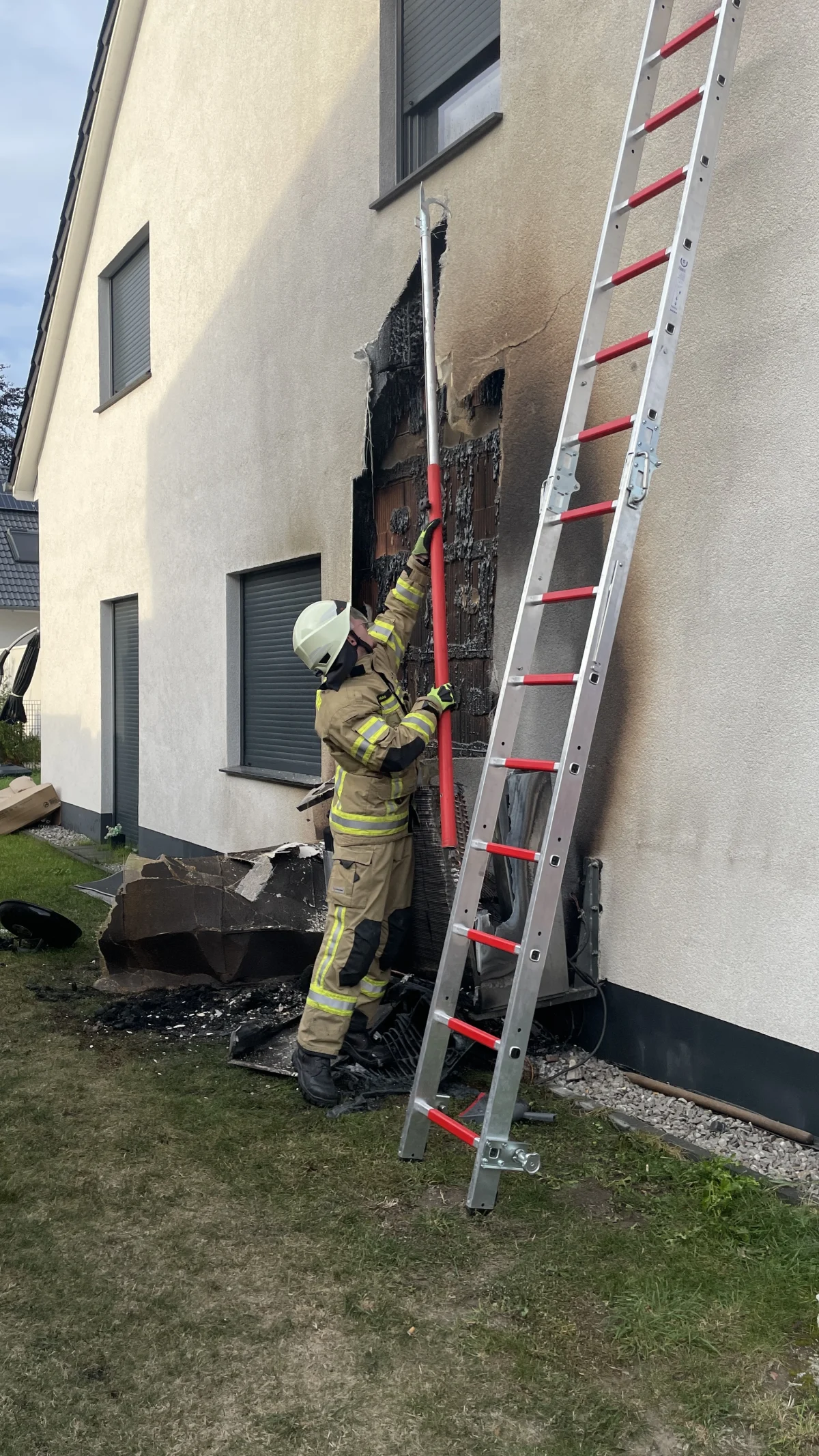 Ein Feuerwehrmann in Einsatzkleidung untersucht die Fassade eines Hauses, die durch einen Brand beschädigt wurde. Eine Leiter lehnt an der Hauswand, und verkohlte Reste sind neben einem Fenster sichtbar.