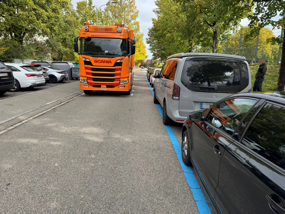 Ein großer orangefarbener Lkw steht frontal auf einer engen Straße. Auf der rechten Seite sind mehrere Autos geparkt, während ein Mann am Straßenrand steht.