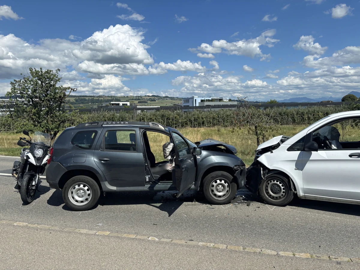 Ein dunkelgraues Auto ist frontal mit einem weißen Fahrzeug kollidiert. Ein Motorrad steht daneben auf der Straße.