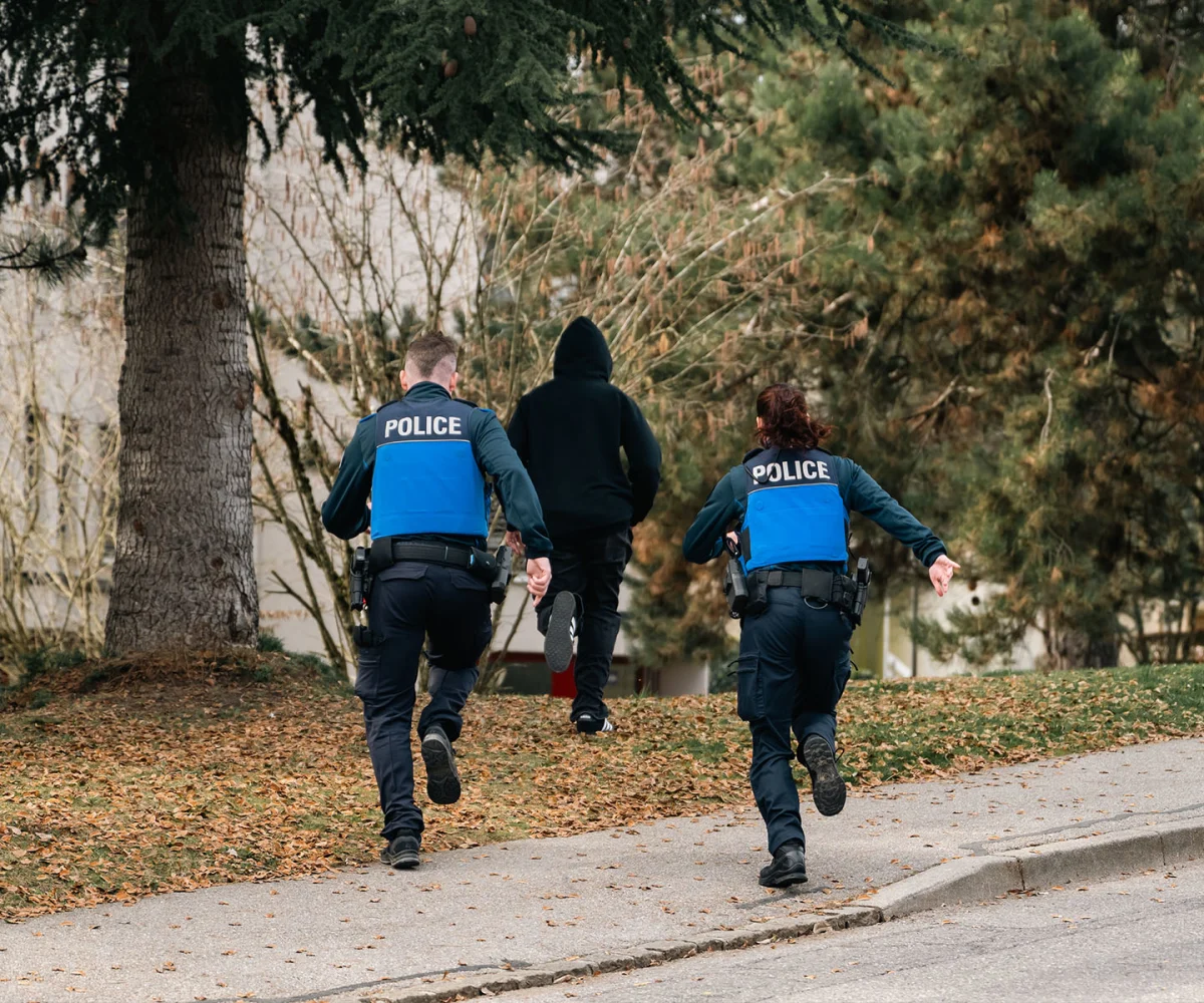 Zwei Polizisten mit blauen Westen und der Aufschrift 'POLICE' jagen eine vermummte Person durch einen baumreichen Park.