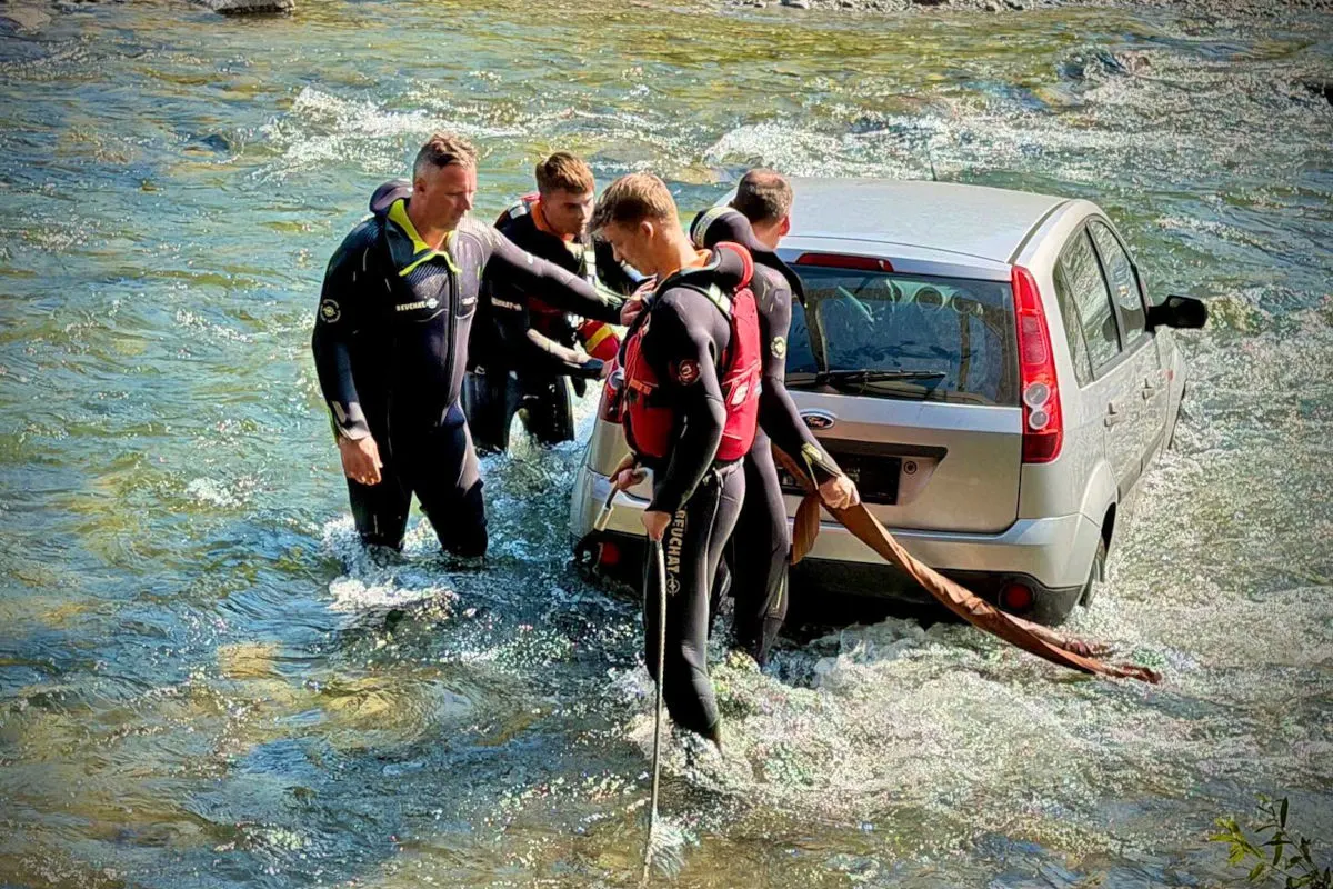 Vier Taucher in Neoprenanzügen stehen in einem Fluss und arbeiten an einem im Wasser stehenden Auto. Sie scheinen es zu sichern oder zu bergen.