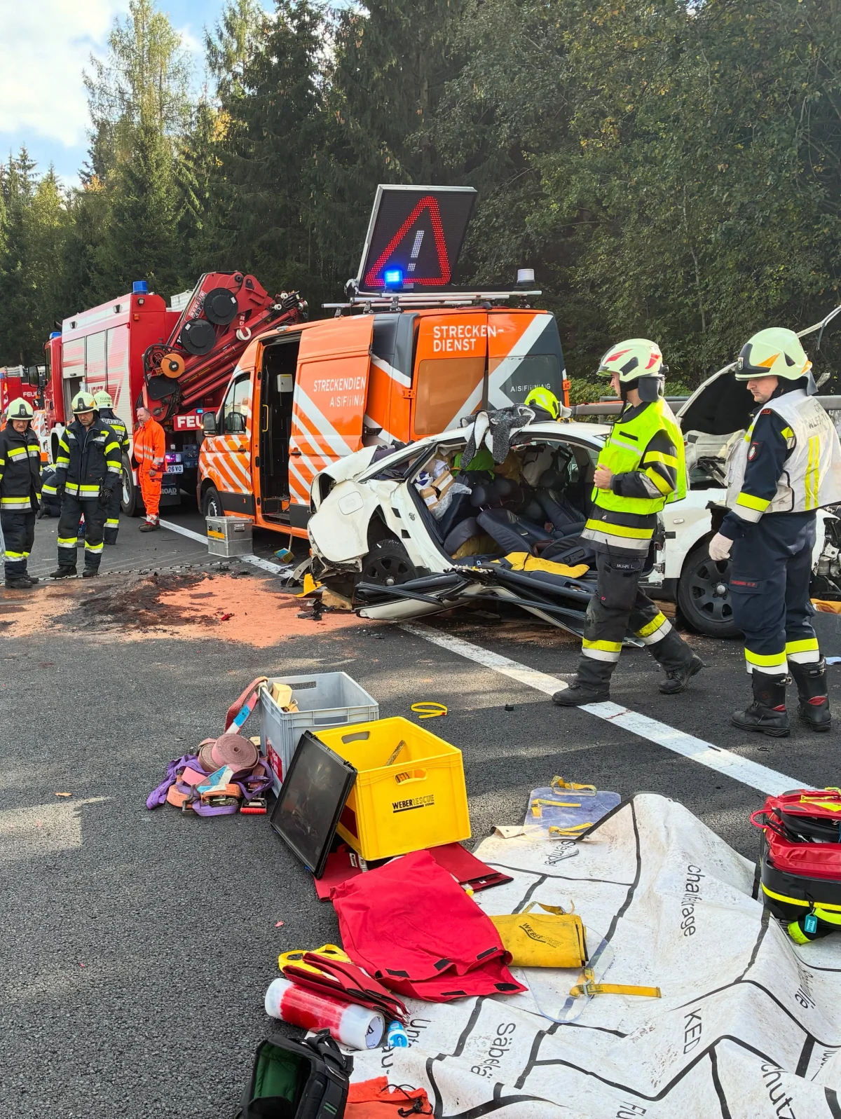 Auf der Autobahn wird ein schwer beschädigtes weißes Auto von Rettungskräften umgeben. Im Hintergrund sind ein Feuerwehrfahrzeug und ein Streckendienst-Fahrzeug zu sehen.
