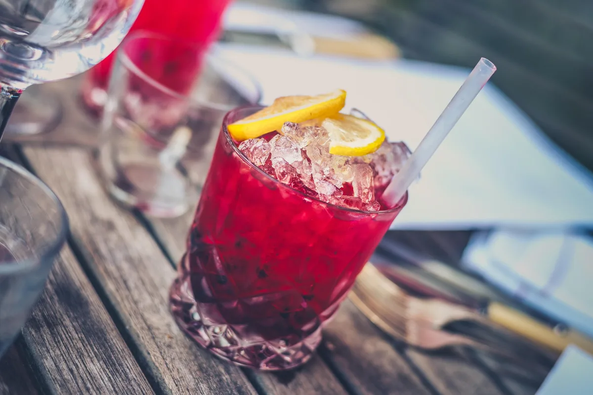Ein Glas mit einem roten, eisgekühlten Getränk, dekoriert mit Zitronenscheiben, steht auf einem Holztisch im Freien.
