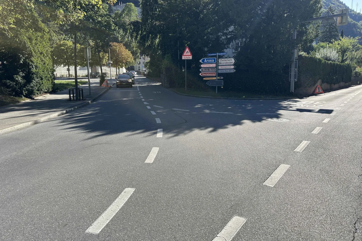 Ein schwarzes Auto fährt auf eine Kreuzung zu, an der mehrere Straßenschilder stehen. Die Szene ist von Bäumen gesäumt und die Sonne scheint.