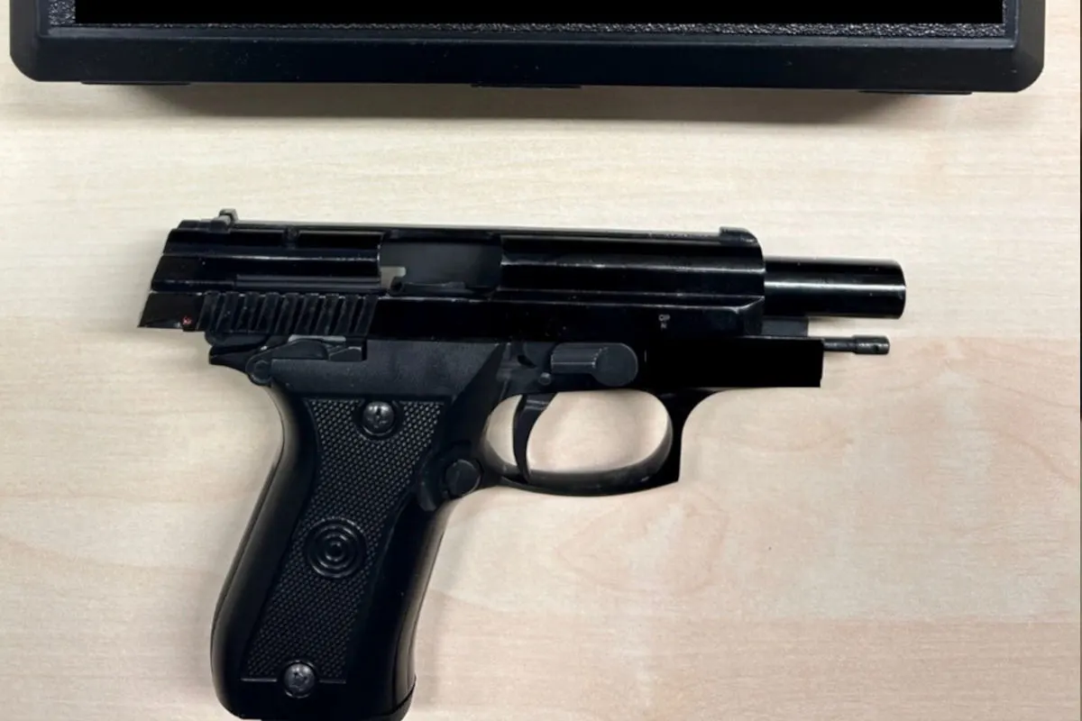 Zu sehen ist eine schwarze Pistole, die flach auf einem hellen Tisch liegt. Oben im Bildrand befindet sich ein schwarzes, rechteckiges Objekt.