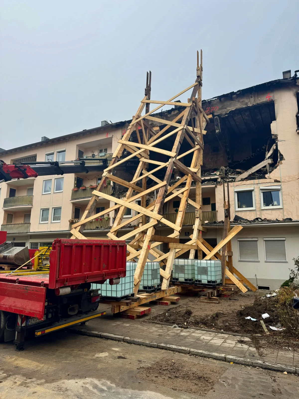 Ein schwer beschädigtes Wohnhaus wird durch umfangreiche Holzkonstruktionen abgestützt. Ein roter Lkw ist im Vordergrund sichtbar.