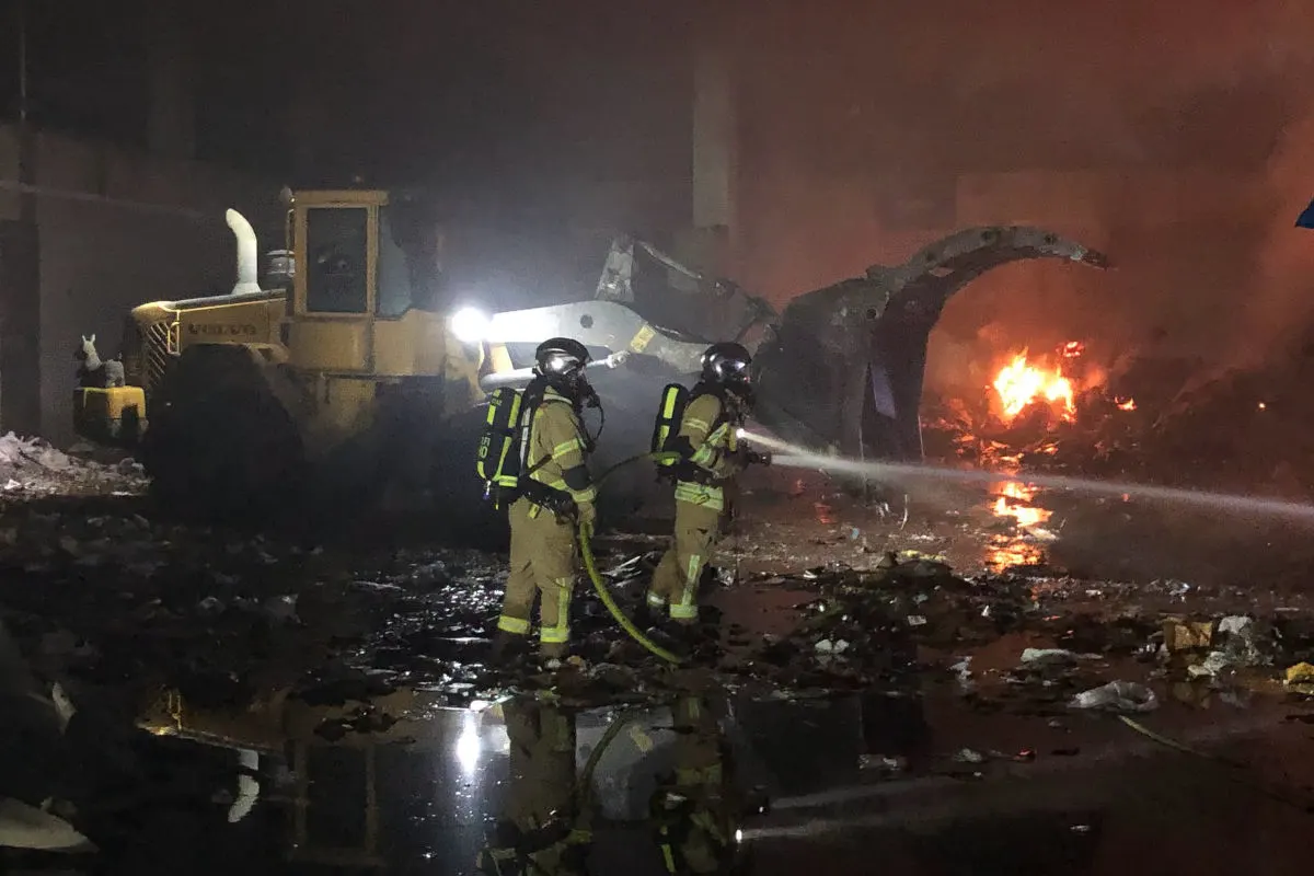 Zwei Feuerwehrleute in Schutzkleidung bekämpfen nachts einen Brand in einer Industriehalle. Ein Bagger steht im Hintergrund, Flammen sind zu sehen.