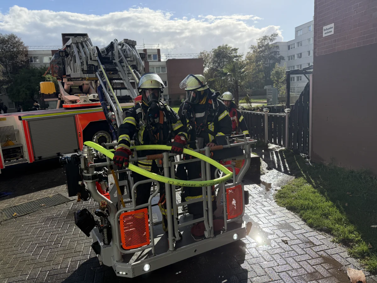 Feuerwehrleute in voller Montur, inklusive Atemschutz, stehen auf einer Drehleiterplattform. Im Hintergrund sind Wohngebäude und ein Feuerwehrfahrzeug sichtbar.