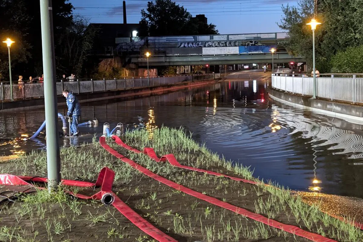 Eine Person steht knietief in einer überfluteten Straße, während rote Feuerwehrschläuche entlang des Ufers verlaufen. Im Hintergrund ist eine Brücke zu sehen, und es ist nachts.