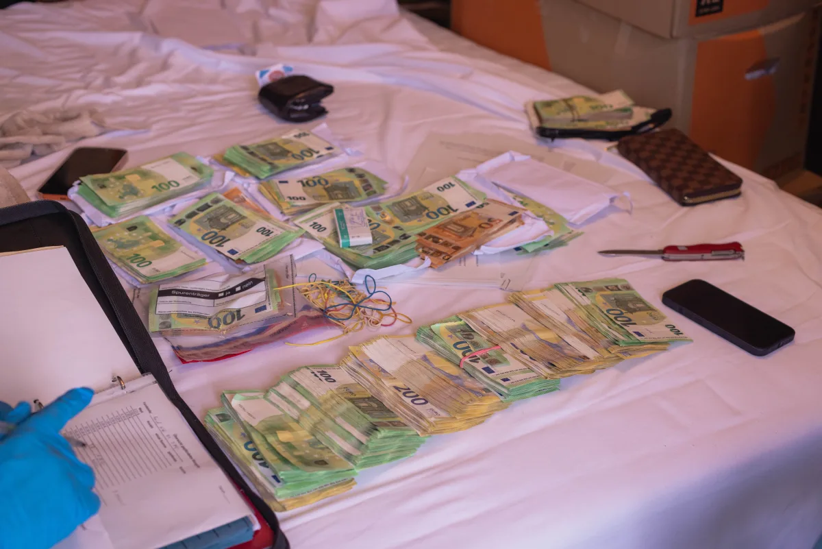 Auf einem Bett liegen zahlreiche Euro-Geldscheine unterschiedlicher Stückelungen, ein Notizbuch, ein Mobiltelefon und ein Messer. Eine Person mit Handschuhen ist am linken Bildrand beim Schreiben zu sehen.