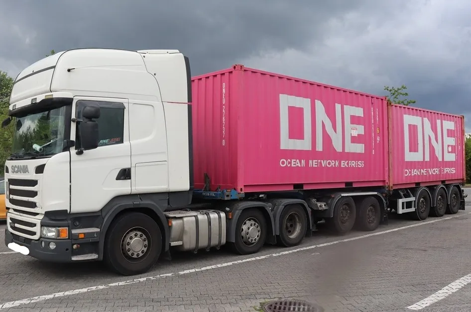 Ein weißer Scania-LKW zieht zwei rosa Container mit der Aufschrift 'ONE' auf einem Parkplatz. Der Himmel ist bewölkt.