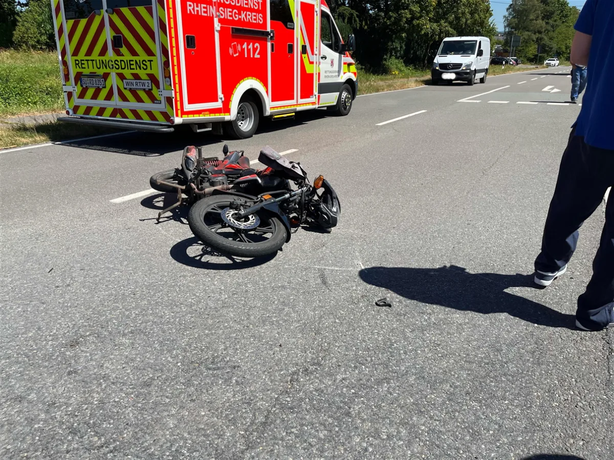 Ein Motorrad liegt umgestürzt auf der Straße. Im Hintergrund steht ein Rettungswagen des Rhein-Sieg-Kreises. Personen sind teilweise sichtbar.