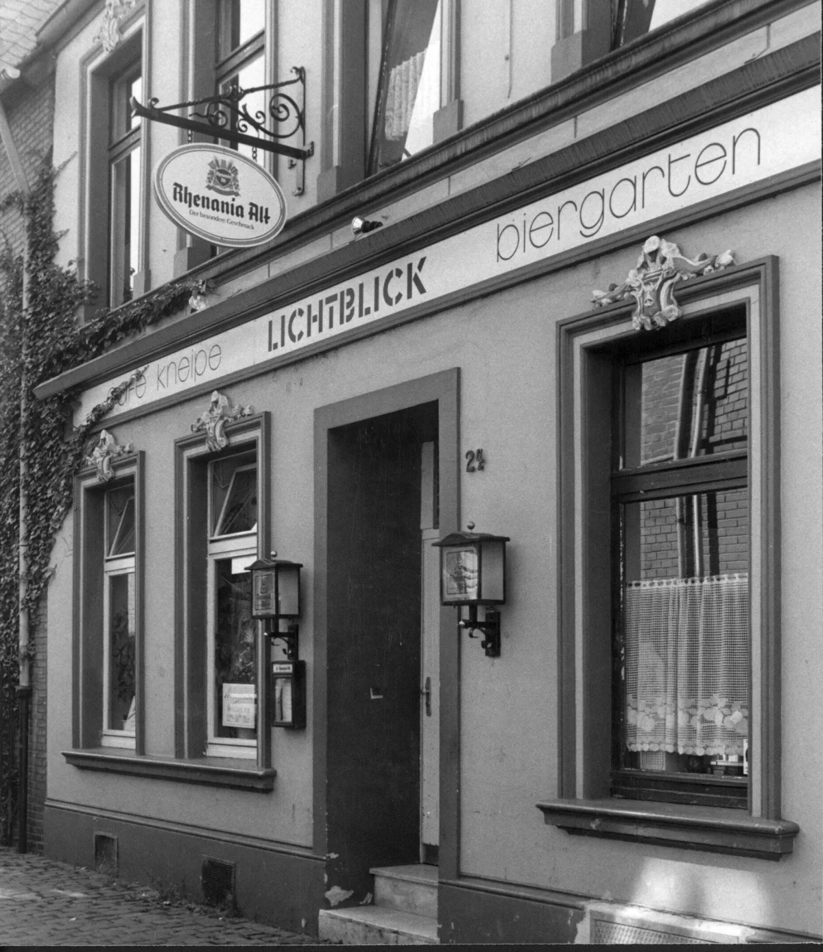 Zu sehen ist der Eingang einer Kneipe mit der Aufschrift 'Lichtblick' und einem Schild 'Rhenania Alt'. An der Fassade sind zwei Fenster und eine Tür erkennbar.