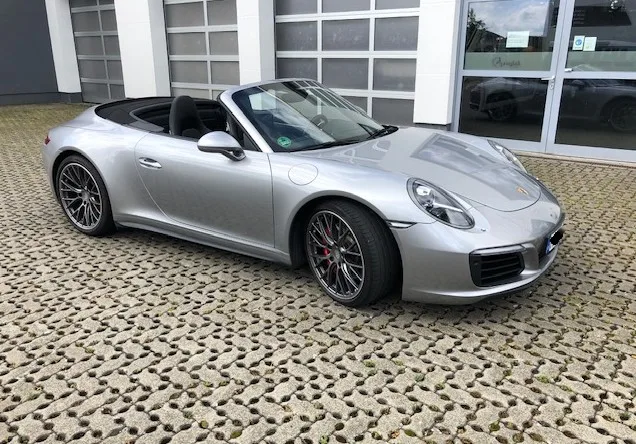 Ein silberner Porsche Cabrio geparkt auf einem gemusterten Pflastersteinparkplatz vor einem modernen Gebäude mit Glasfront.