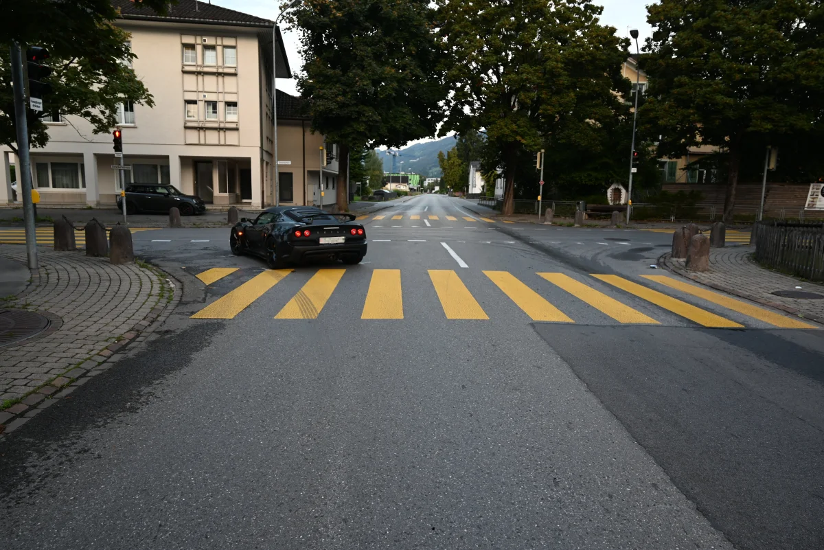 Ein schwarzer Sportwagen fährt über einen leeren Zebrastreifen, während die Ampel rot zeigt. Im Hintergrund sind Gebäude und Bäume zu sehen.