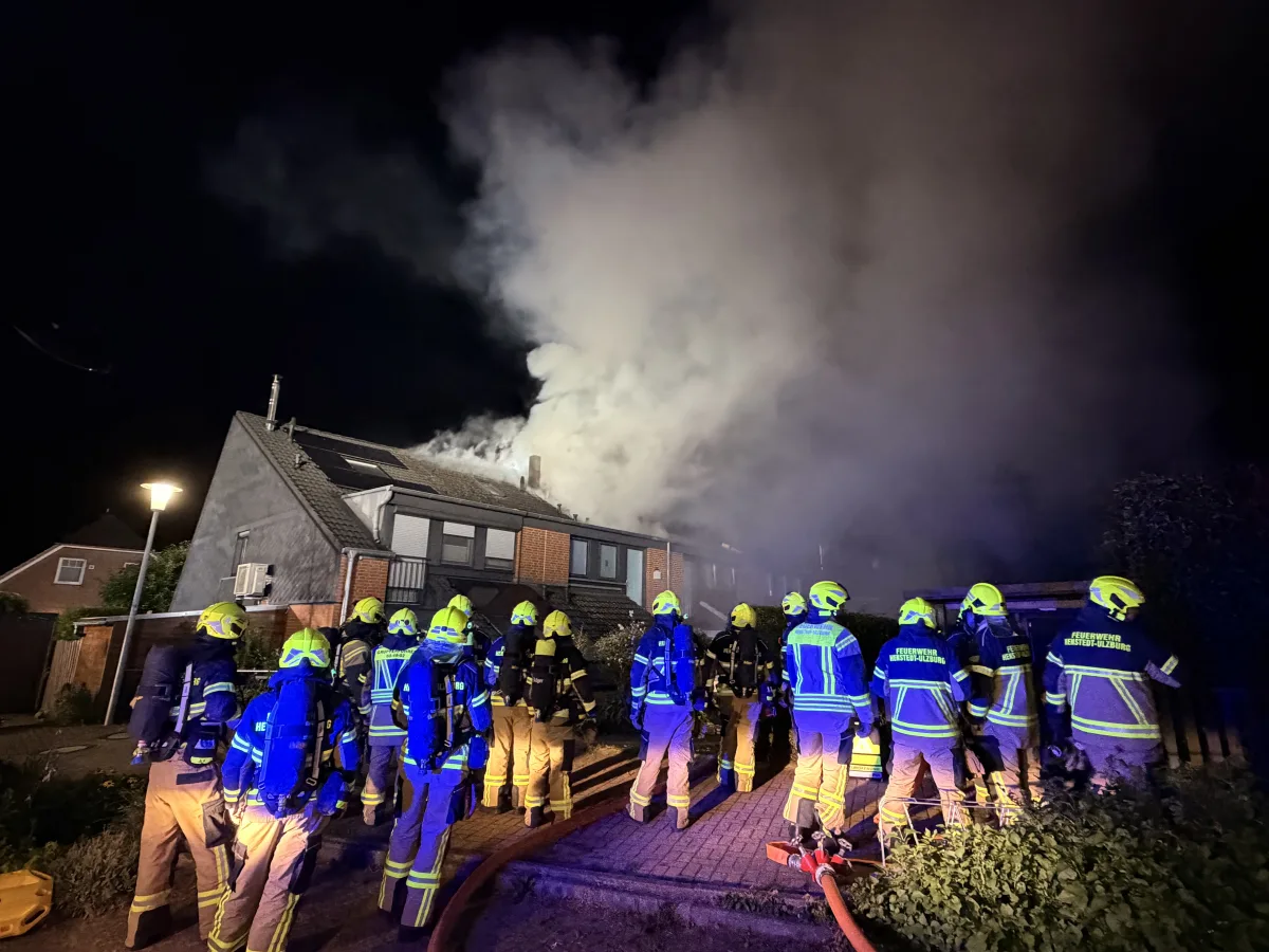 Mehrere Feuerwehrleute in voller Ausrüstung stehen vor einem Haus, dessen Dach brennt. Dunkler Rauch steigt in die Nacht auf.
