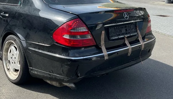 Ein schwarzer Mercedes mit sichtbaren Beschädigungen. Die Heckklappe ist mit Gurten befestigt.