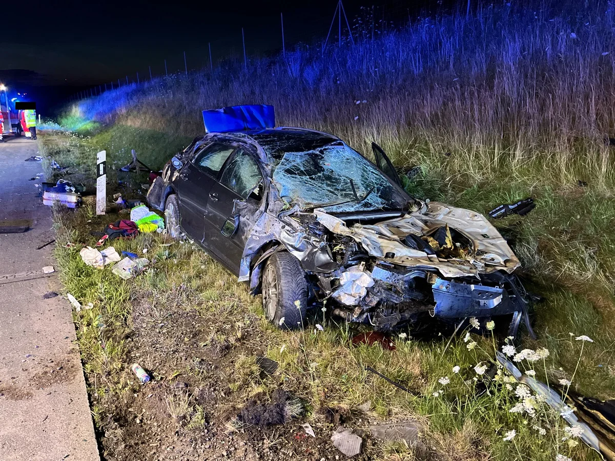 Ein stark beschädigtes Fahrzeug liegt an einer Böschung neben der Straße. Trümmer sind um das Auto verstreut, und im Hintergrund sind Blaulichter zu sehen.