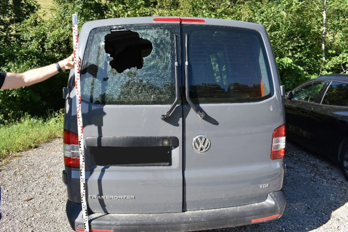 Ein grauer VW Transporter steht auf einem Parkplatz, das Heckfenster ist zersplittert. Links hält jemand einen Maßstab.