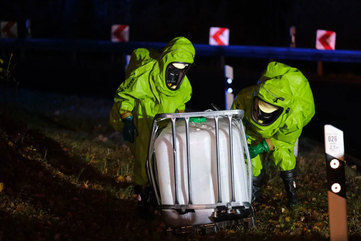 Zwei Personen tragen grüne Schutzanzüge und arbeiten bei Nacht an einem weißen Tank neben einer Landstraße. Der Hintergrund zeigt Verkehrsschilder und eine Leitplanke.