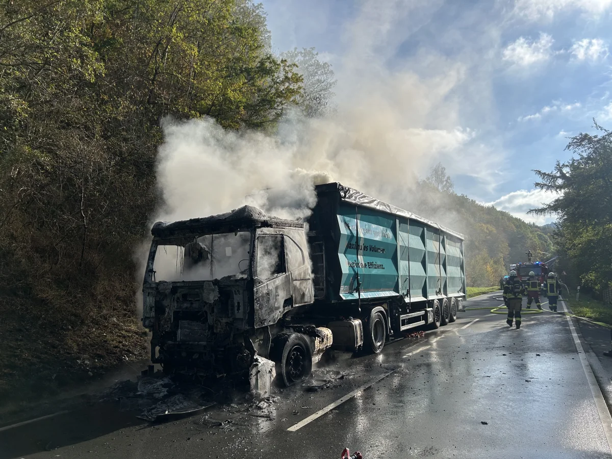 Ein ausgebrannter LKW steht auf einer Landstraße, umgeben von Rauch. Feuerwehrleute sind anwesend.