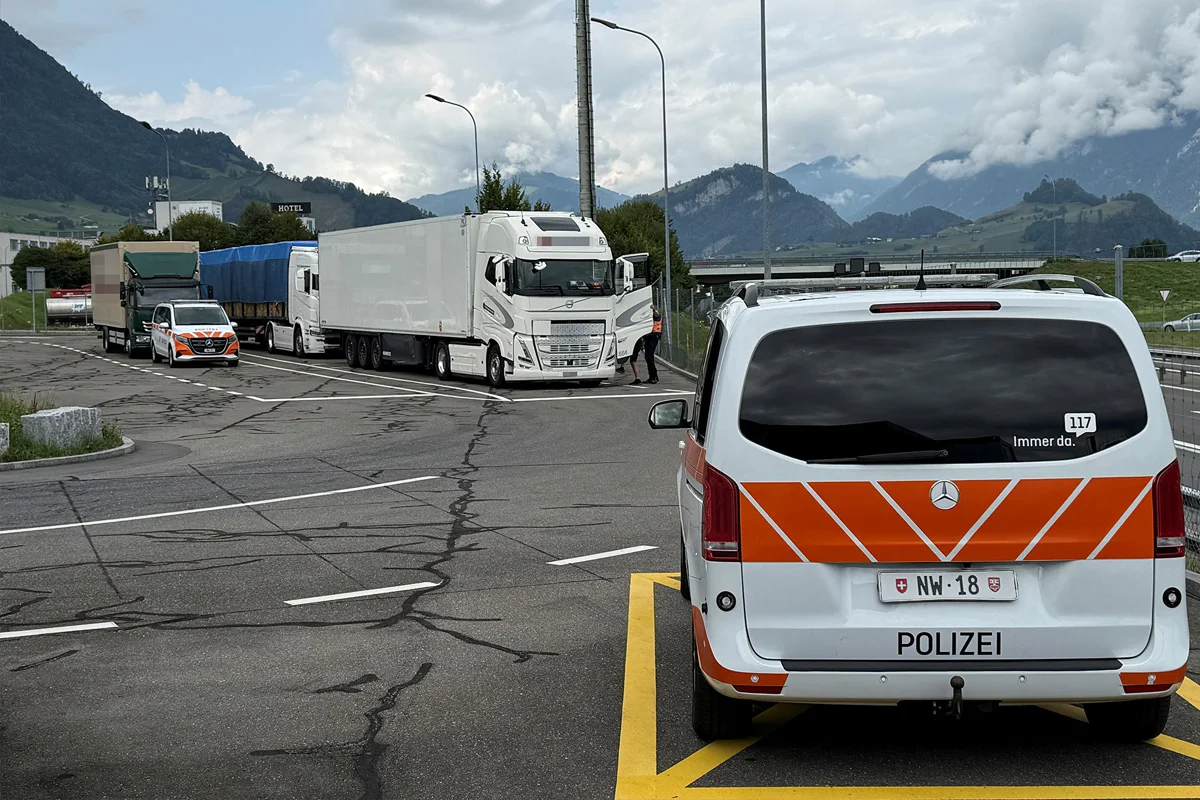 Ein Polizeiauto steht auf einem markierten Parkplatz im Vordergrund, während mehrere Lkw im Hintergrund parken. Die Szene hat eine bergige Landschaft mit bewölktem Himmel.