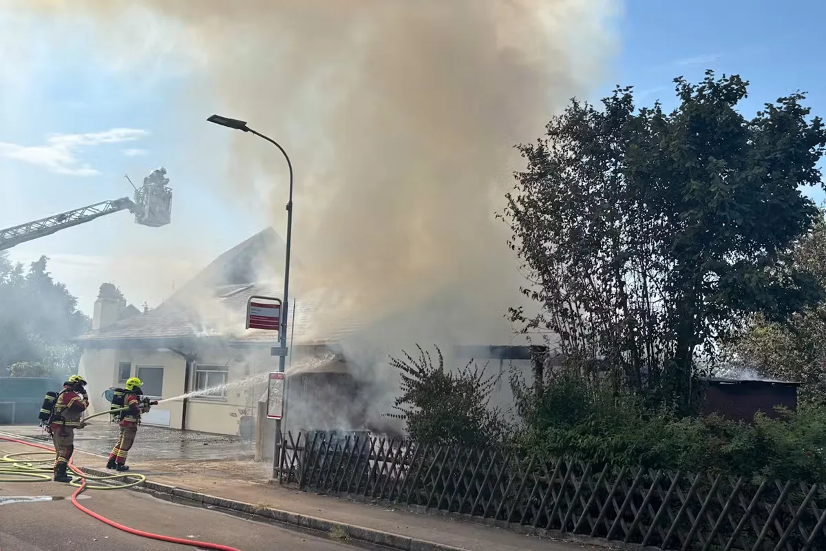 Zwei Feuerwehrleute bekämpfen einen Brand an einem Wohnhaus; dichter Rauch steigt auf.