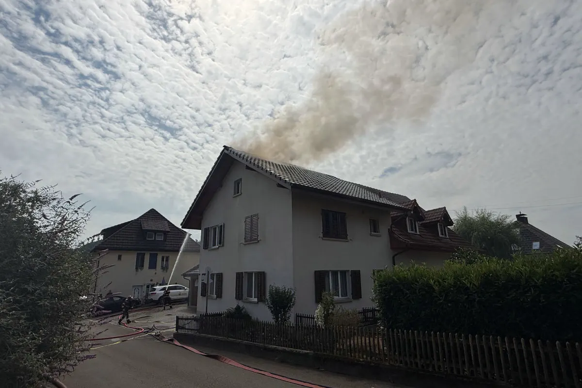 Ein zweistöckiges Wohnhaus mit Rauch, der vom Dach aufsteigt. Feuerwehrleute sind mit Löschschläuchen im Einsatz.
