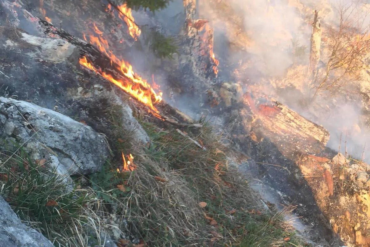 Flammen lodern an einem steilen Hang, durchmischt mit Gras, Felsen und leichtem Rauch. Der Brand scheint sich auszubreiten.