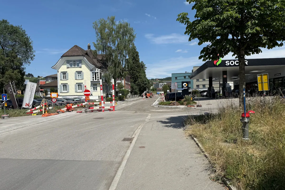 Eine Baustelle mit Absperrungen und verschiedenen Verkehrsschildern befindet sich vor einem Wohngebiet. Im Hintergrund ist eine Tankstelle sichtbar.
